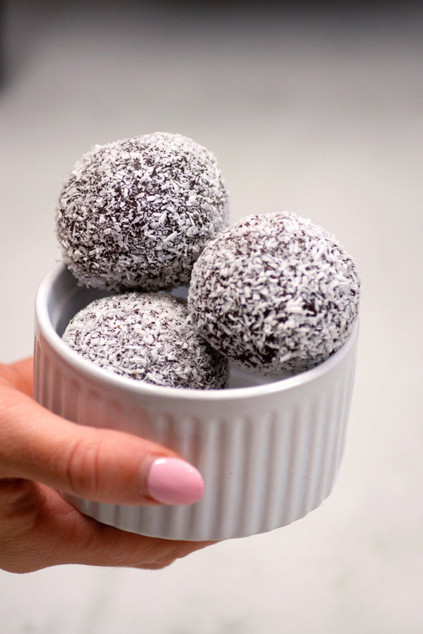 Hand holding a small white bowl with three grayish-brown balls, possibly truffles or cookies, on a light gray background.